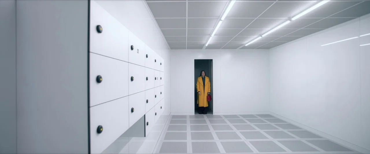The Substance (2024) movie still: wide — A stark white, clinical room with grid-patterned floor tiles and uniform metal locker banks. A woman in a…
