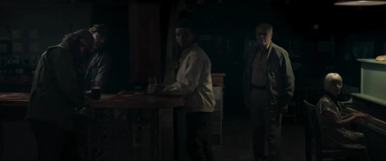 Offseason (2022) movie still: wide — A dimly lit bar with a wooden counter and a piano against the wall. Four people occupy the bar area while a…