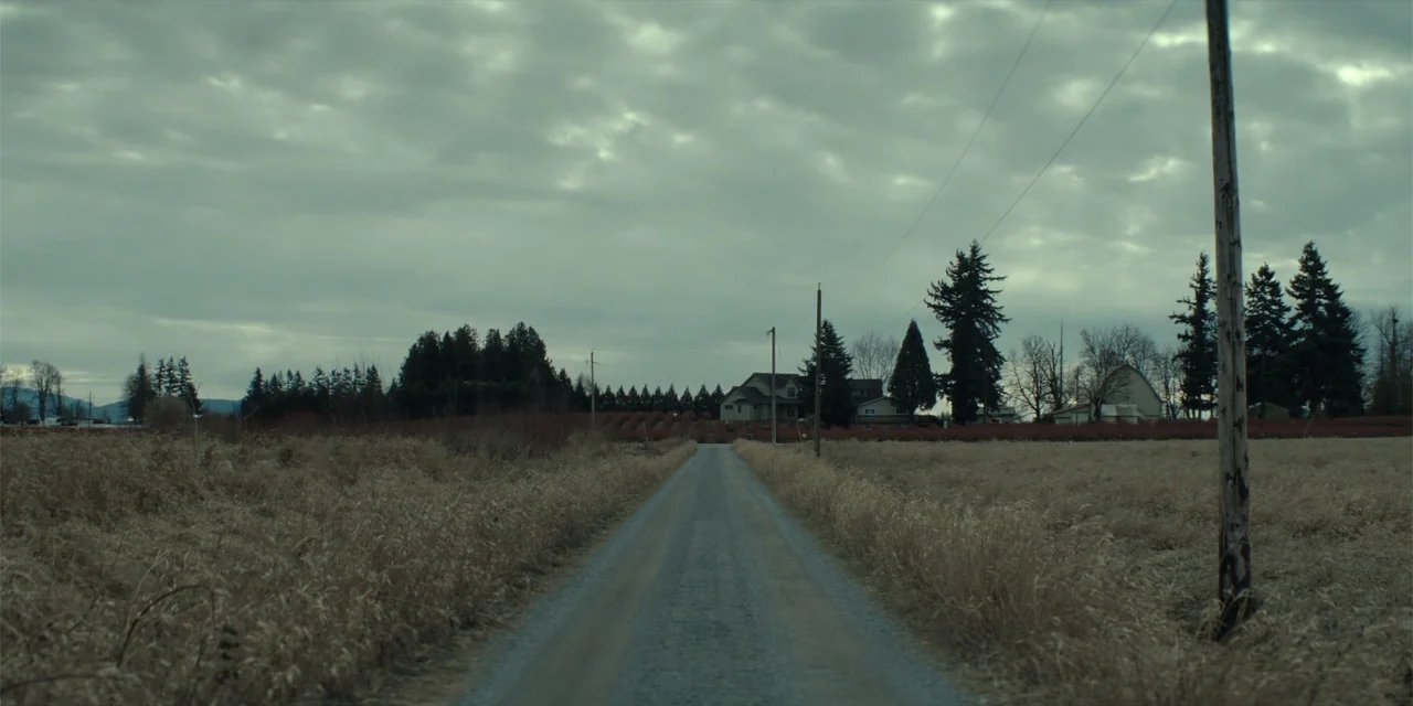 The Monkey (2025) movie still: establishing — A gravel road cuts through dry fields towards distant farmhouses under a cloudy sky. The landscape…