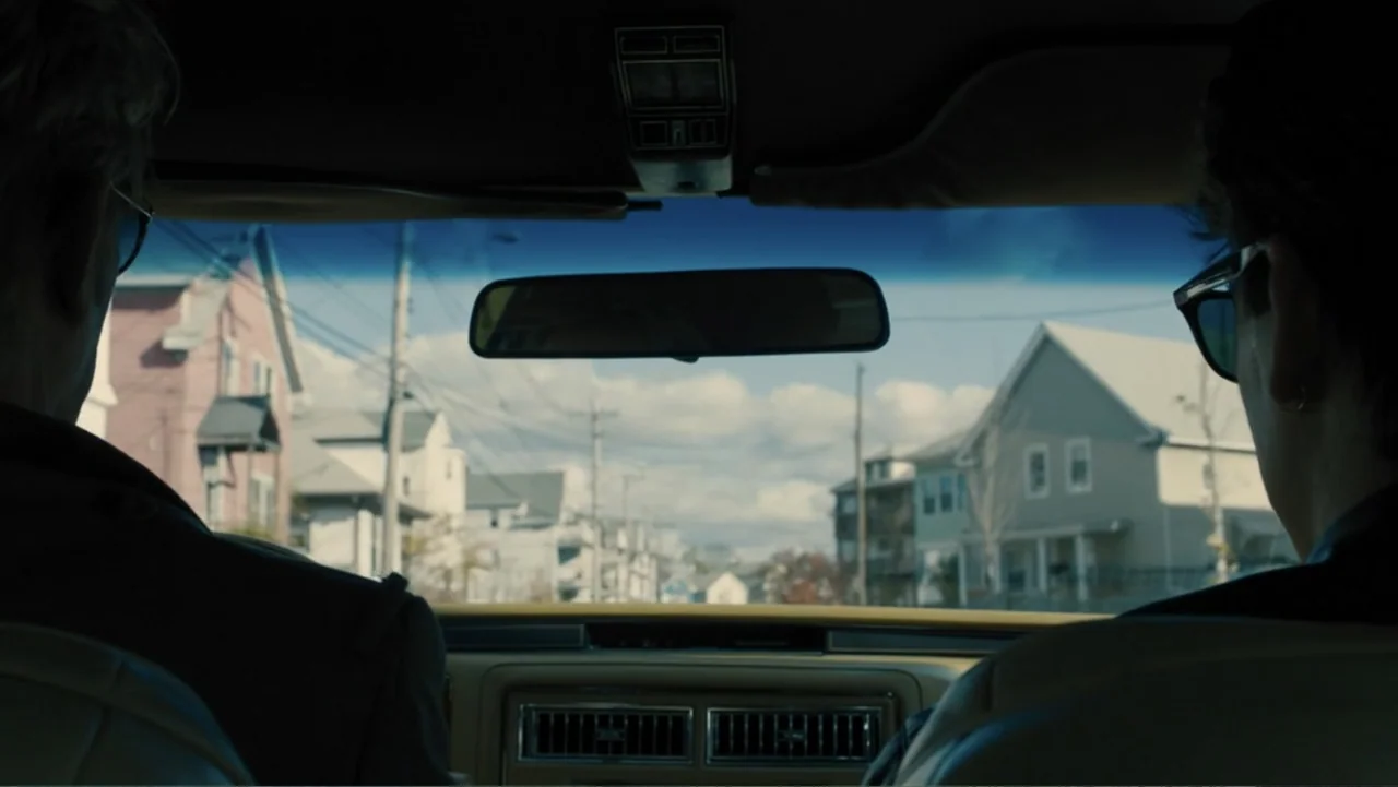Bleed for This (2016) movie still: medium — A view from inside a car looking out at a quiet residential suburban street. Two men sit in the front…