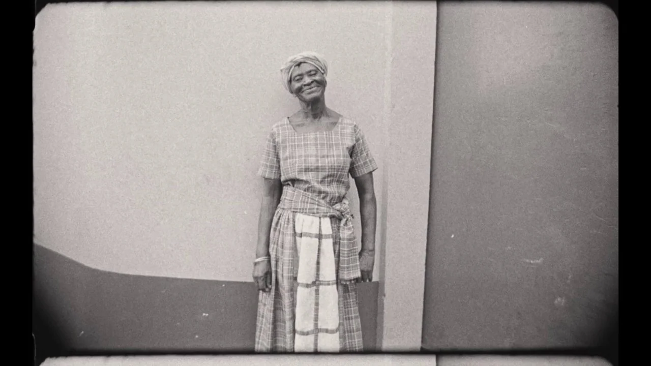 Black Mother (2018) movie still: fullbody — An elderly woman stands in the corner of a simple concrete exterior structure. She wears a plaid dress…