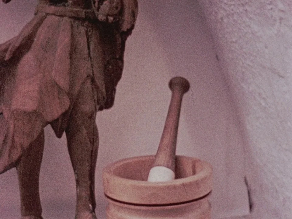 The Wormwood Star (1956) movie still: closeup — A wooden figurine stands next to a wooden mortar and pestle. The textured surface of a wall appears…