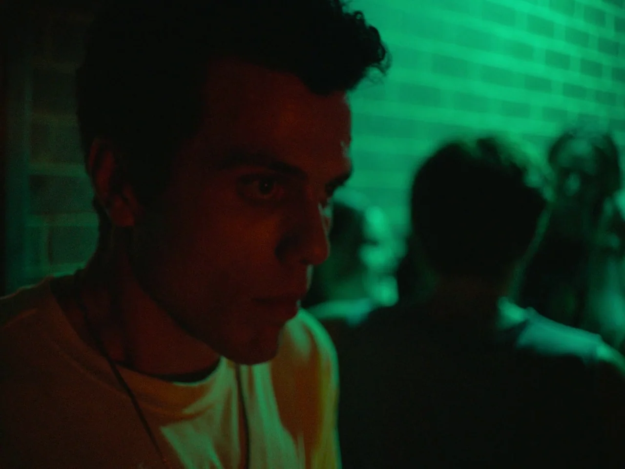 Of An Age (2022) movie still: closeup — A dark, dimly lit environment features a brick wall illuminated by a green light. A young man with dark wavy…