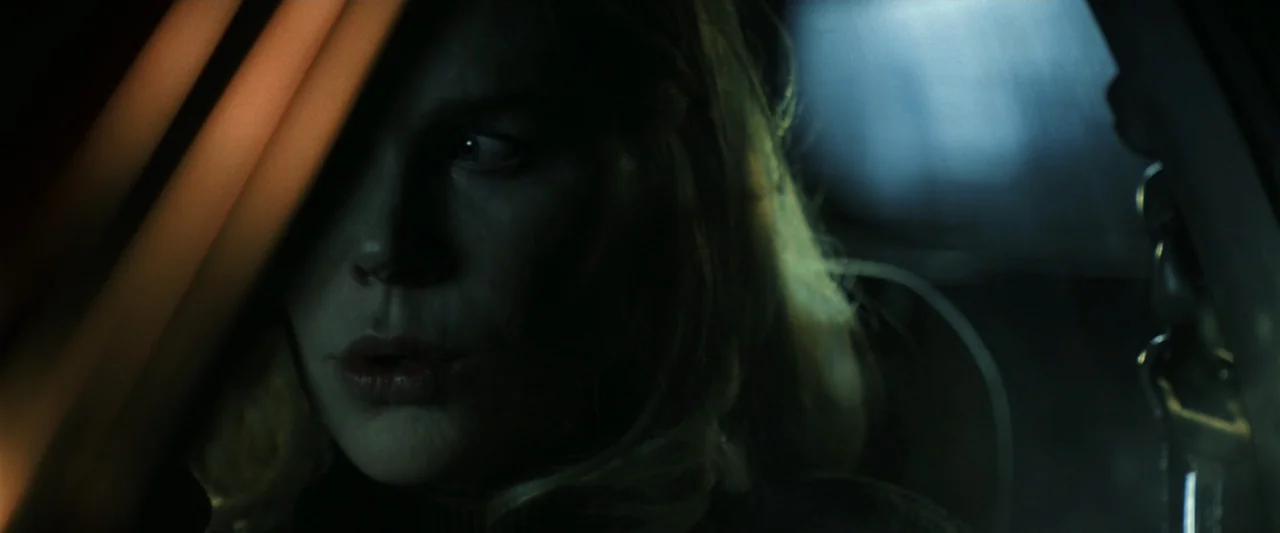 Holland (2025) movie still: closeup — The interior of a vehicle at night with low ambient light sources. A woman sits in the driver seat looking…