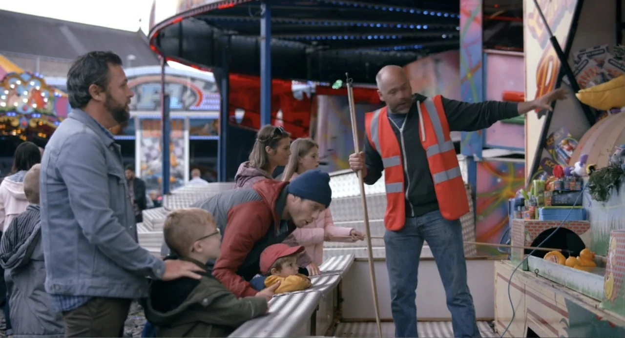 Living Out Loud (1998) movie still: medium — A bustling outdoor carnival area features a colorful game booth with yellow rubber ducks. A worker in…