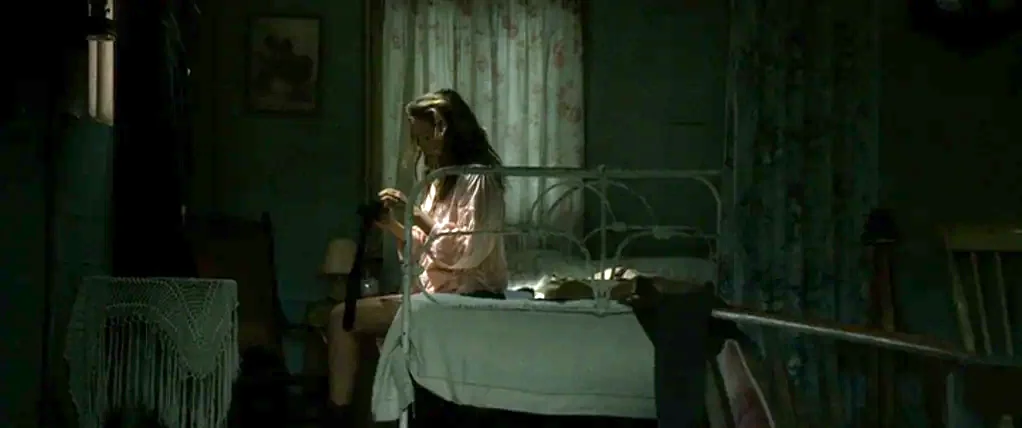 Ondine (2009) movie still: medium — A dimly lit bedroom features an iron-framed bed and floral curtains. A woman in a pink top sits on the mattress…