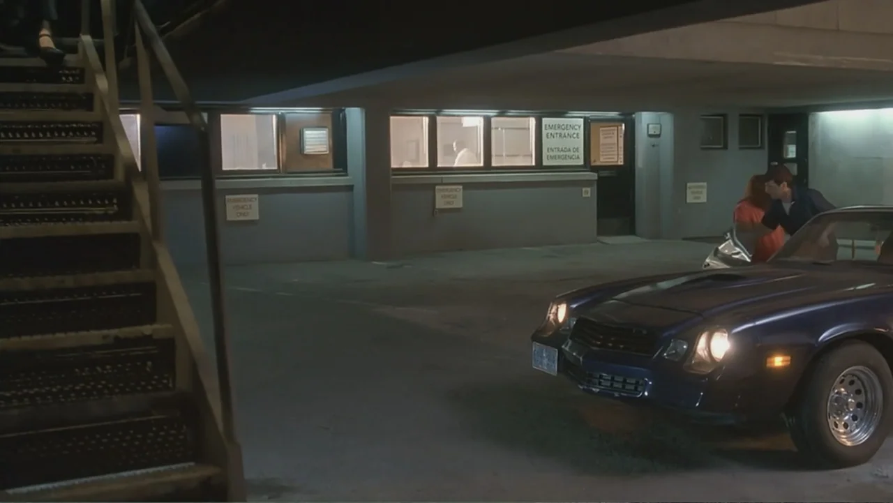 The Art of War (2000) movie still: wide — A dimly lit hospital emergency entrance area is framed by a metal staircase. Two people step out of a dark…