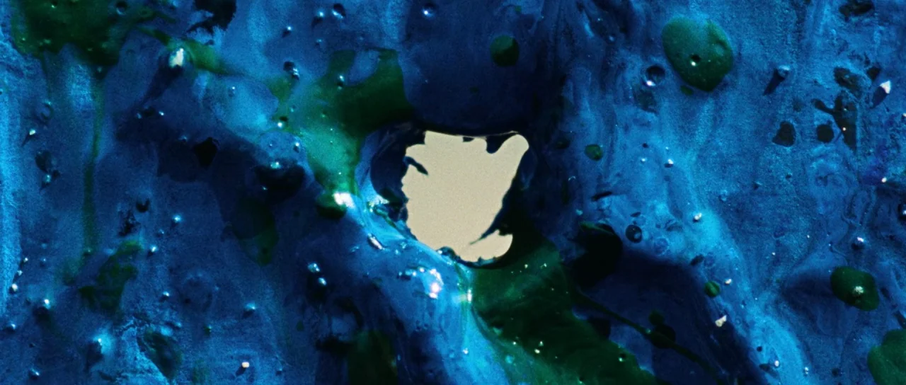 Let The Corpses Tan (2017) movie still: closeup — A thick layer of glossy, viscous blue and green paint covers a surface. An irregular hole in the…
