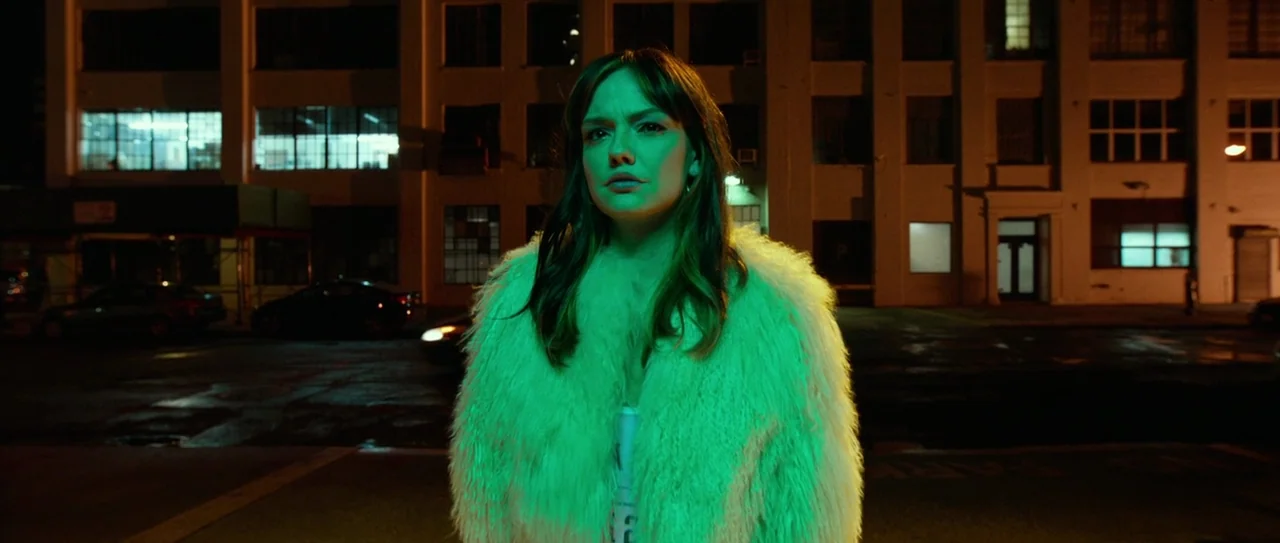 Nerve (2016) movie still: medium — A woman stands in a dimly lit parking lot in front of a multi-story building. Harsh green light casts a strong…