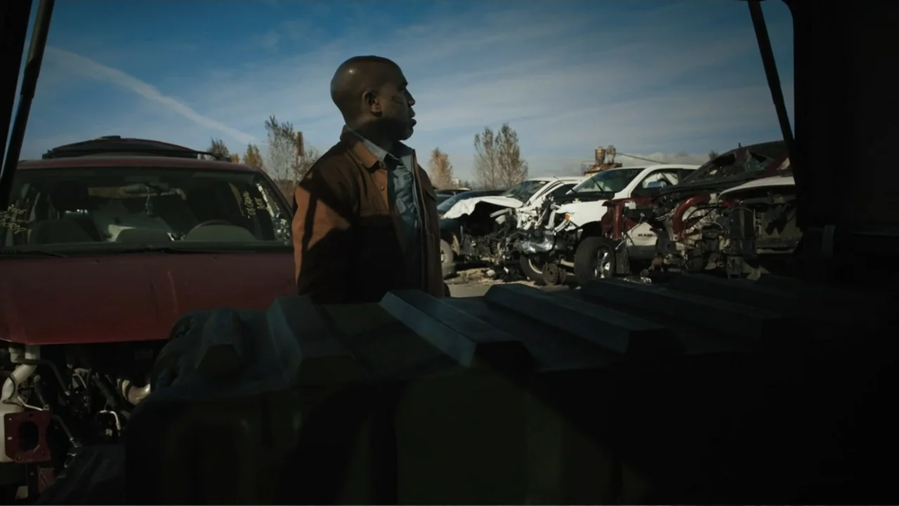 Bullet Proof (2022) movie still: medium — An outdoor car graveyard filled with wrecked vehicles under a clear blue sky. A man in a brown jacket…