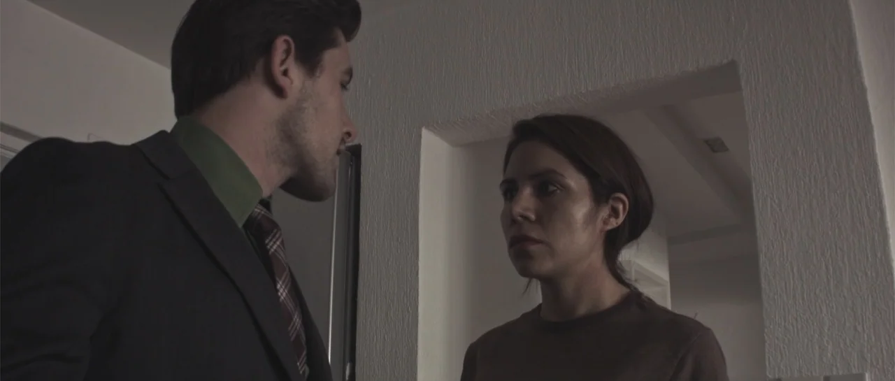 Amor Mutante (2019) movie still: medium — A minimalist white apartment interior features a doorway and stark white textured walls. A man in a dark…