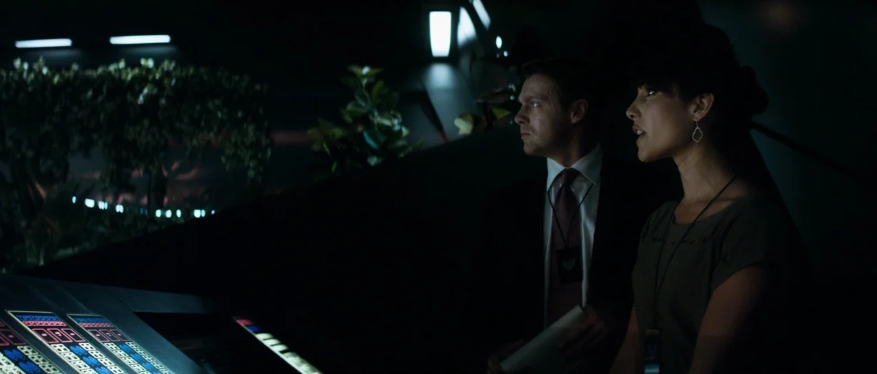 Elysium (2013) movie still: medium — A dimly lit high-tech control center features rows of illuminated consoles and potted plants. A man and a woman…