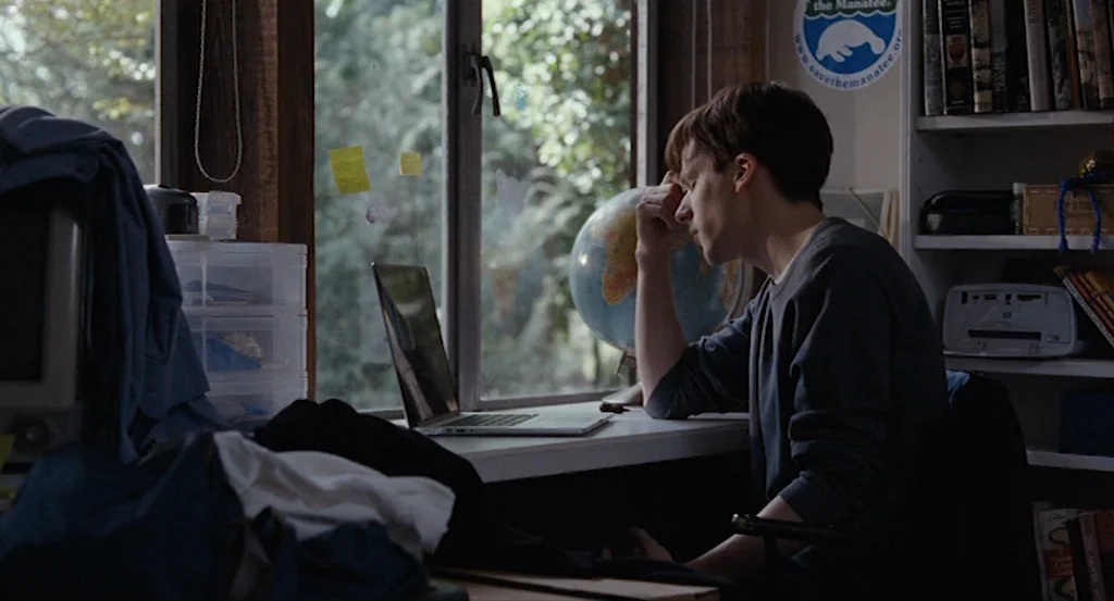 Louder Than Bombs (2015) movie still: medium — A cluttered room features a wooden desk set against a large window with greenery. A young man sits in…