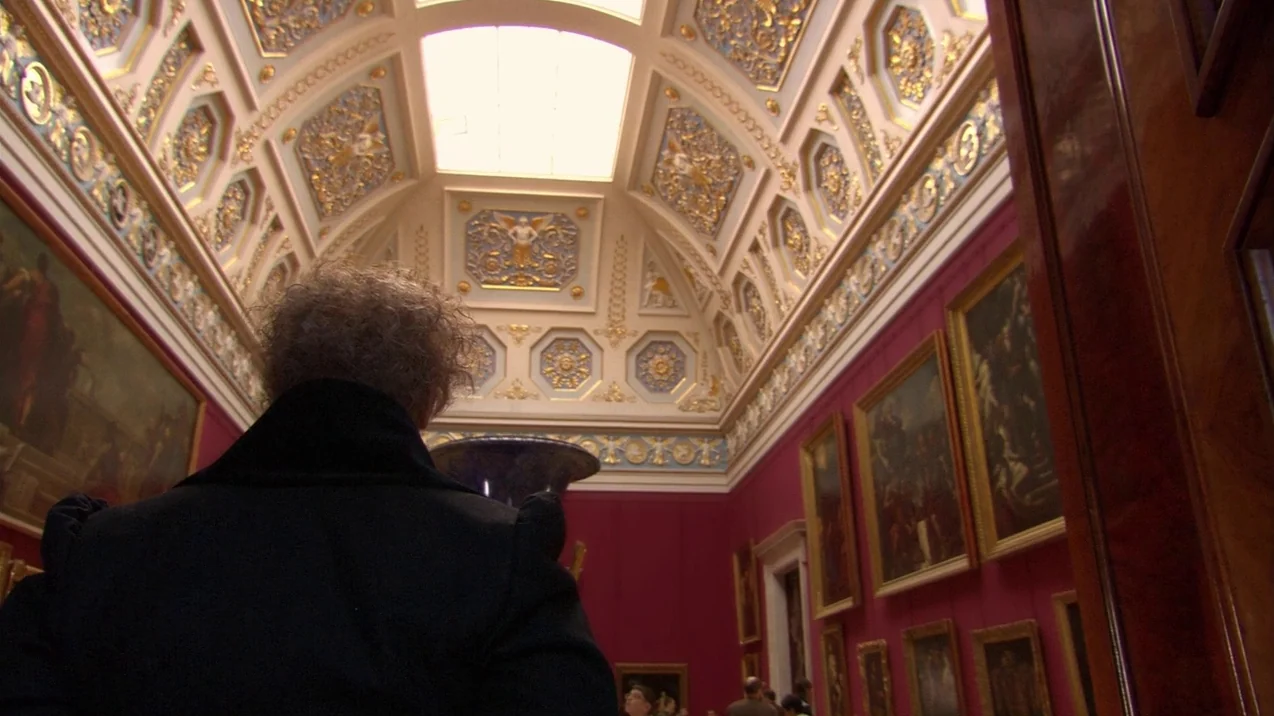 Russian Ark (2002) movie still: medium — A grand gallery room features deep red walls adorned with framed oil paintings. A person seen from behind…