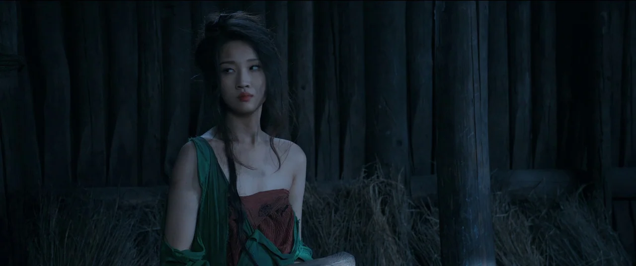Full River Red (2023) movie still: medium — A dimly lit room features a backdrop of vertical wooden logs and gathered straw. A woman wearing green…