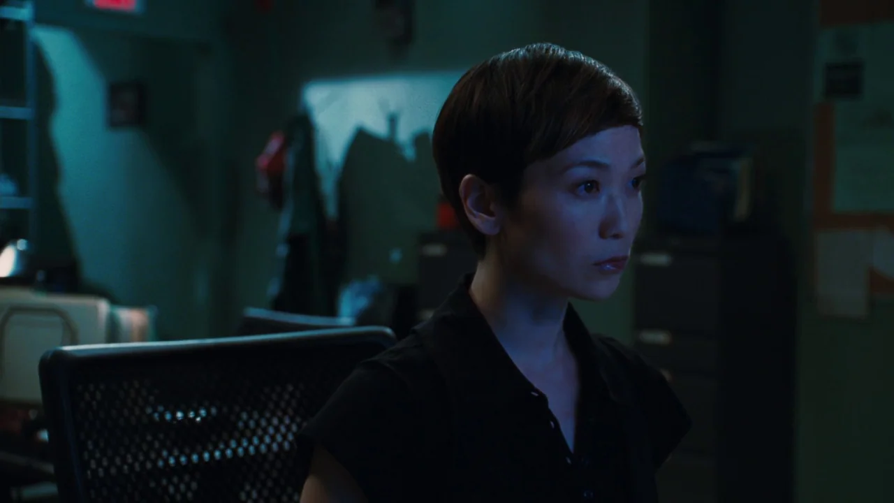 Saw VI movie still: medium — A dimly lit modern office space with dark walls and a blurred workstation background. A woman with short hair sits in a…