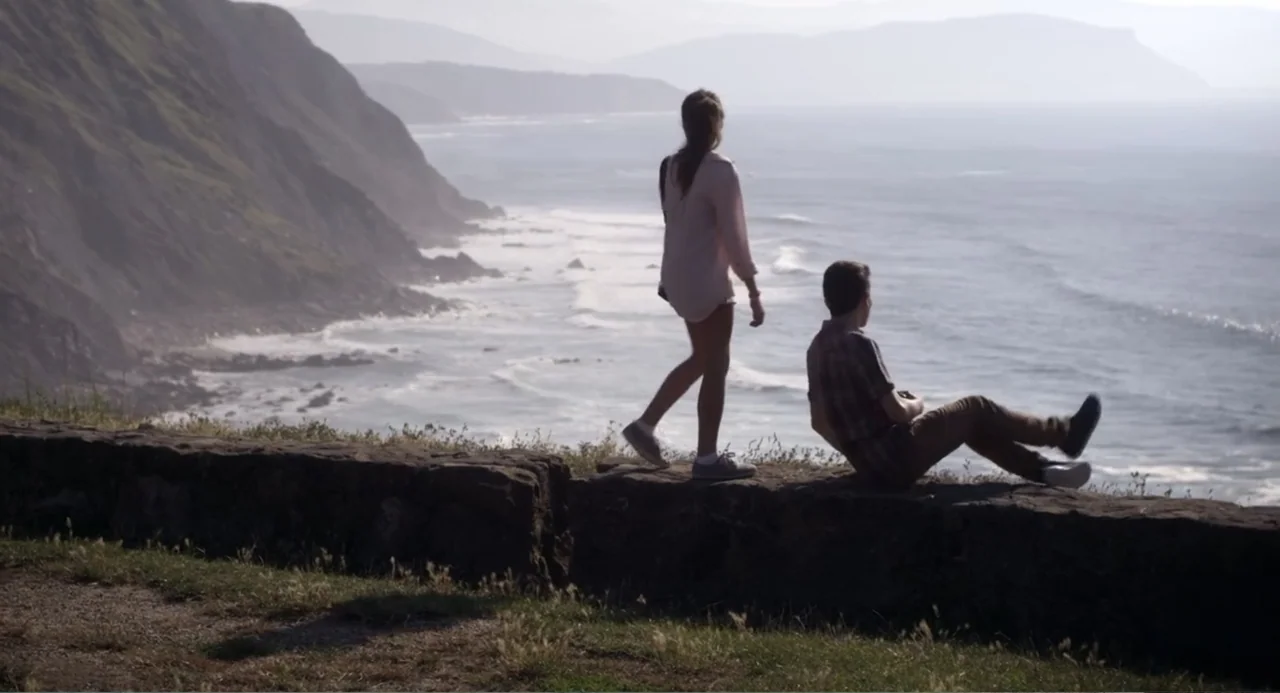 Newton's Third Law (2017) movie still: fullbody — A rugged cliff edge overlooks a vast ocean with distant hills on the horizon. A woman walks toward…