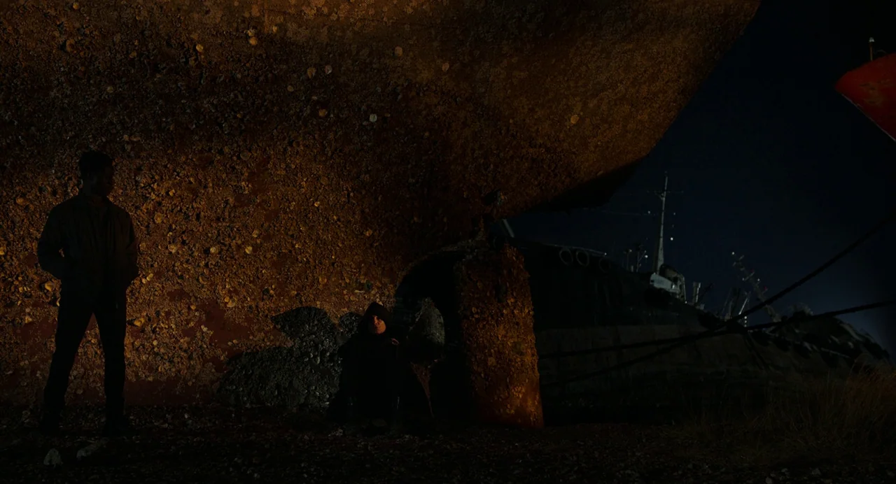 Crimes of the Future (2022) movie still: wide — A massive, rusty ship hull covered in barnacles looms over a dark, gravelly shipyard. Two men stand…
