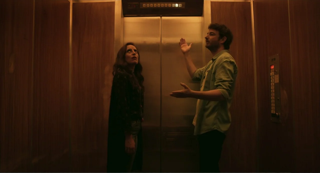 The Lift (2021) movie still: medium — A small elevator interior features wood-paneled walls and a metal control panel button display. A man and a…