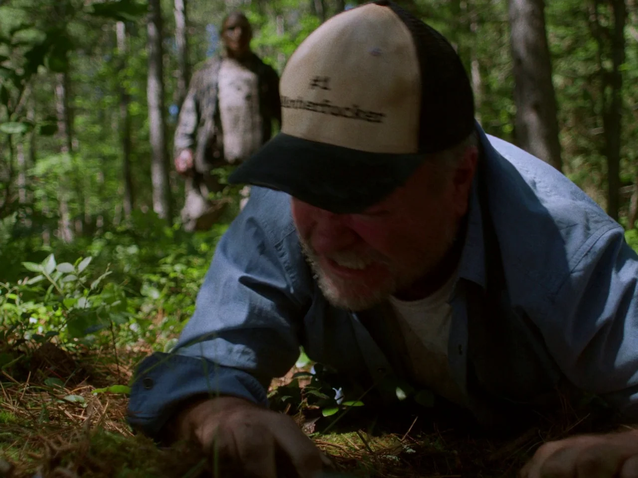 In A Violent Nature (2024) movie still: medium — A dense woodland area with green foliage and pine needles covering the ground surface. A man crawls…