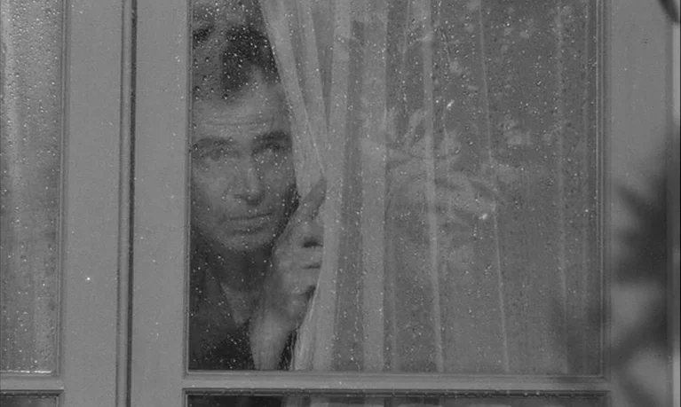 Lolita (1962) movie still: closeup — A rain-streaked window pane separates the interior from the exterior environment. A man peeks through the…