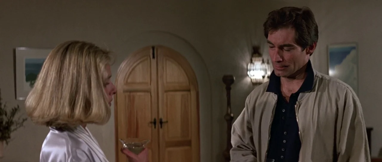 The Living Daylights (1987) movie still: medium — A hotel room with light-colored walls and a distinctive arched wooden double door. A woman in a…