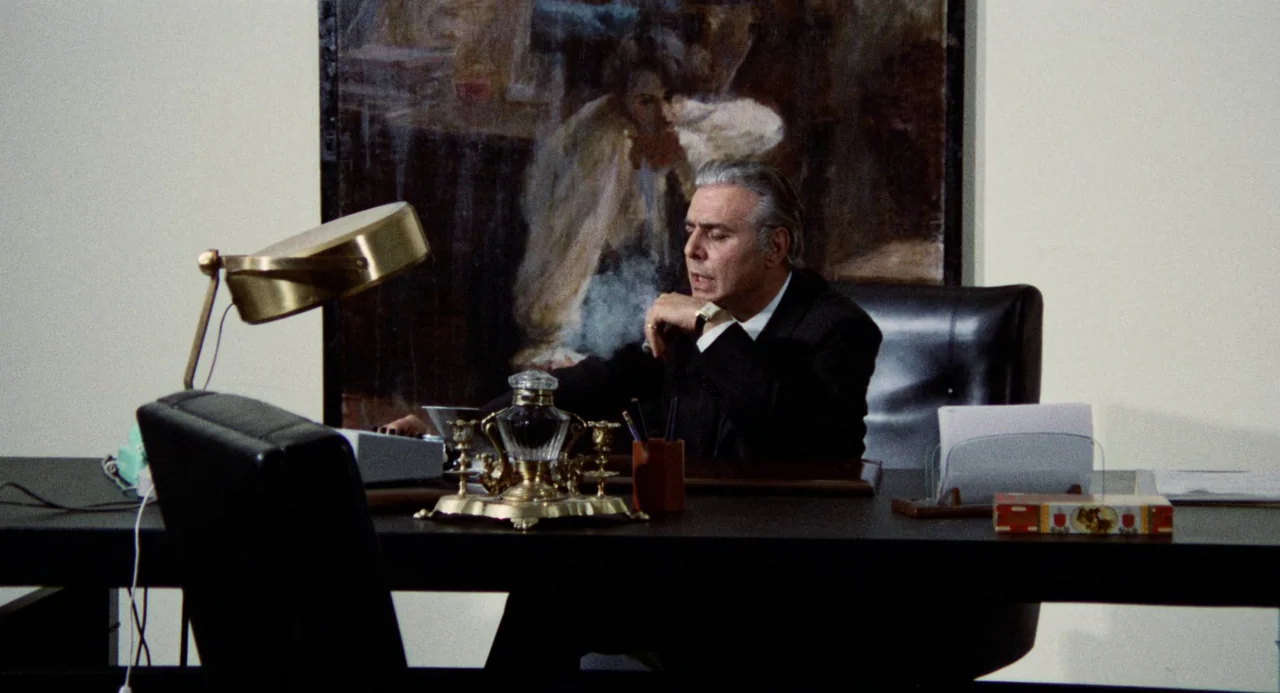 Investigation of a Citizen Above Suspicion (1970) movie still: medium — A man sits behind a dark mahogany desk featuring an ornate inkwell set. He…
