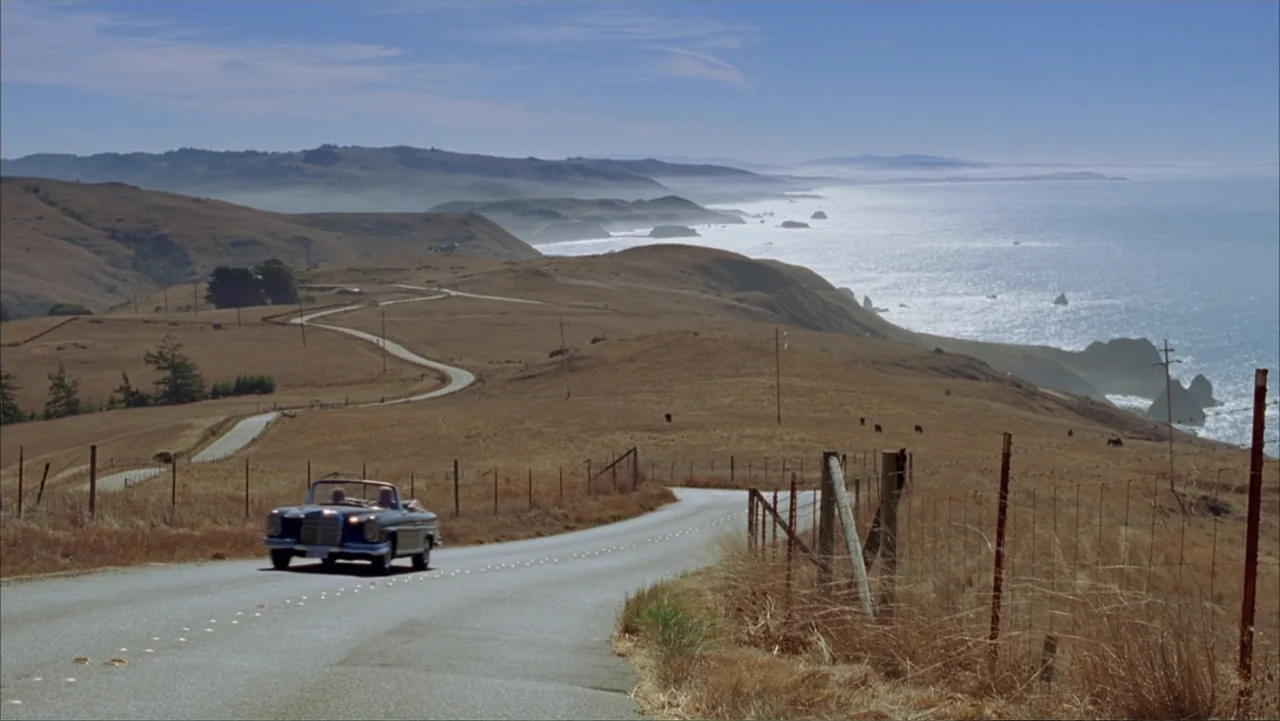 Rumor Has It... (2005) movie still: wide — A winding asphalt road hugs the rolling brown coastal hills along the Pacific Ocean. A dark-colored…