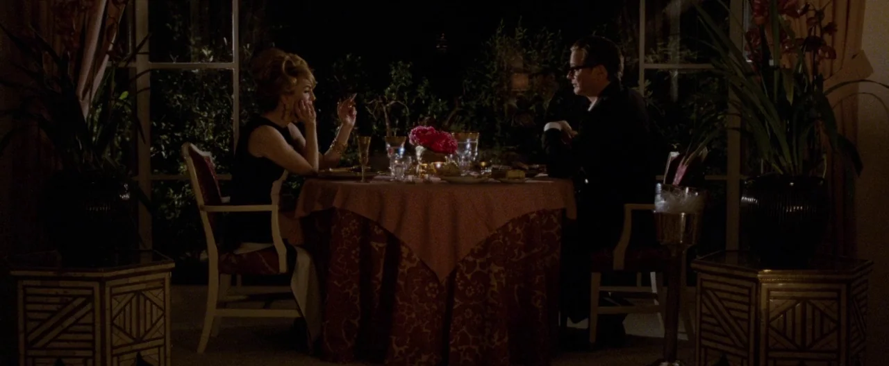 A Single Man (2009) movie still: wide — A dark restaurant interior at night with a round table draped in patterned fabric. A couple sits facing each…