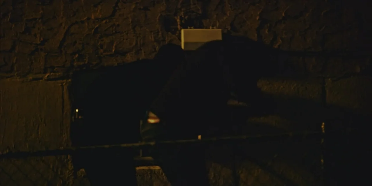 Equal Standard (2021) movie still: medium — An exterior masonry wall at night features a metal chain-link fence in foreground. A dark-clad figure…