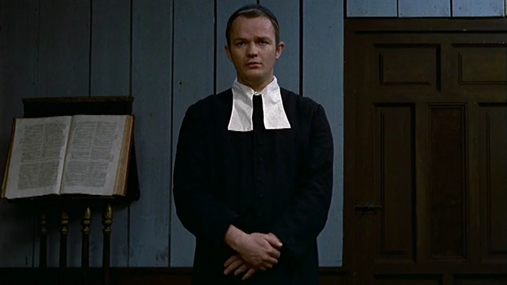 The Scarlet Letter (1973) movie still: medium — A austere room features vertical wooden wall paneling and a dark wooden door. A man in clerical…