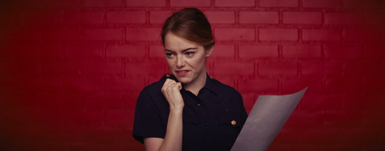 La La Land (2016) movie still: medium — A woman stands before a vibrant red brick wall that fills the frame. She wears a dark blue uniform and holds…
