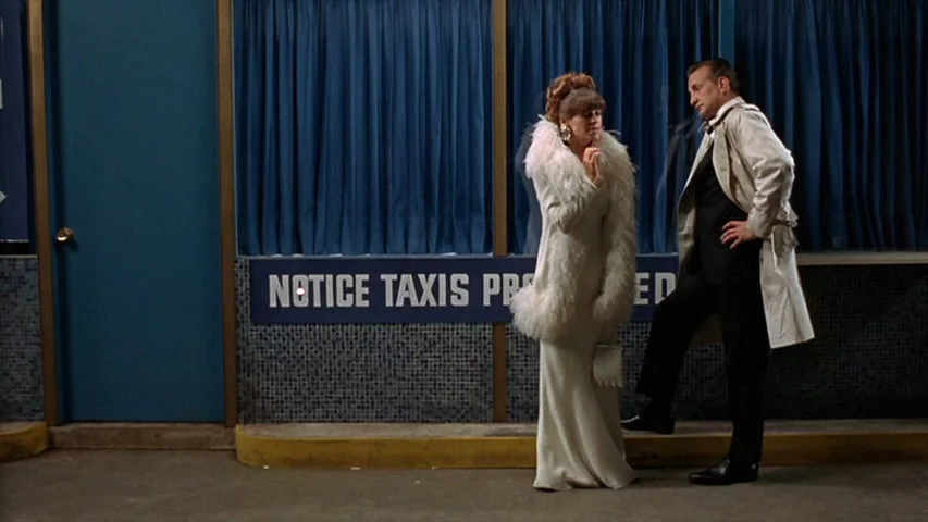 Petulia (1968) movie still: fullbody — A taxi stand features a tiled wall with blue curtains and a signage placard. A woman in a feather-trimmed…