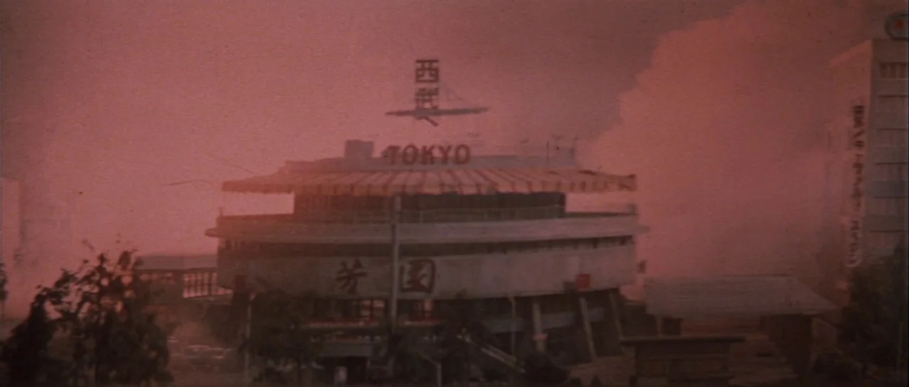 Godzilla Vs Gigan (1972) movie still: establishing — A low-rise circular building with Japanese signage is engulfed in heavy reddish smoke. The…