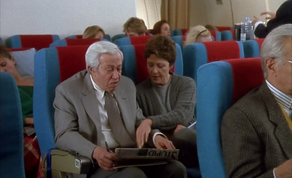 I Want To Go Home (1989) movie still: medium — An airplane cabin features rows of blue seats with vibrant red center panels. A man and woman sit…