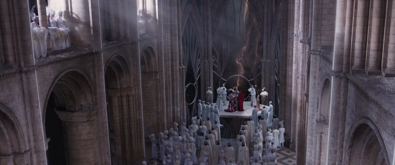 Jupiter Ascending (2015) movie still: wide — A high-angle view shows a stone nave with tall arched walls and vaulted ceilings. A large crowd in…