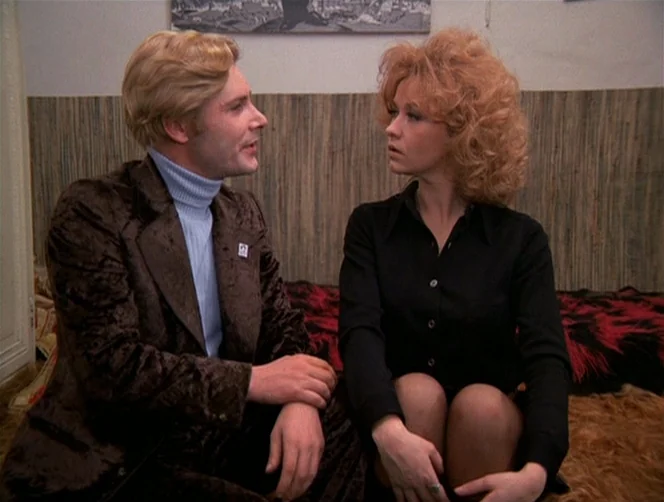 WR: Mysteries of the Organism (1971) movie still: medium — A man and woman sit on a bed in a room with wood paneling. They are facing each other in…