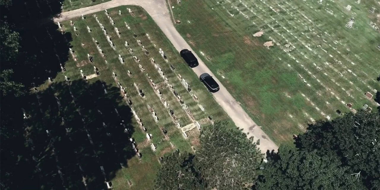 Equal Standard (2021) movie still: establishing — An aerial view of a green cemetery filled with rows of stone markers. Two black cars follow a…