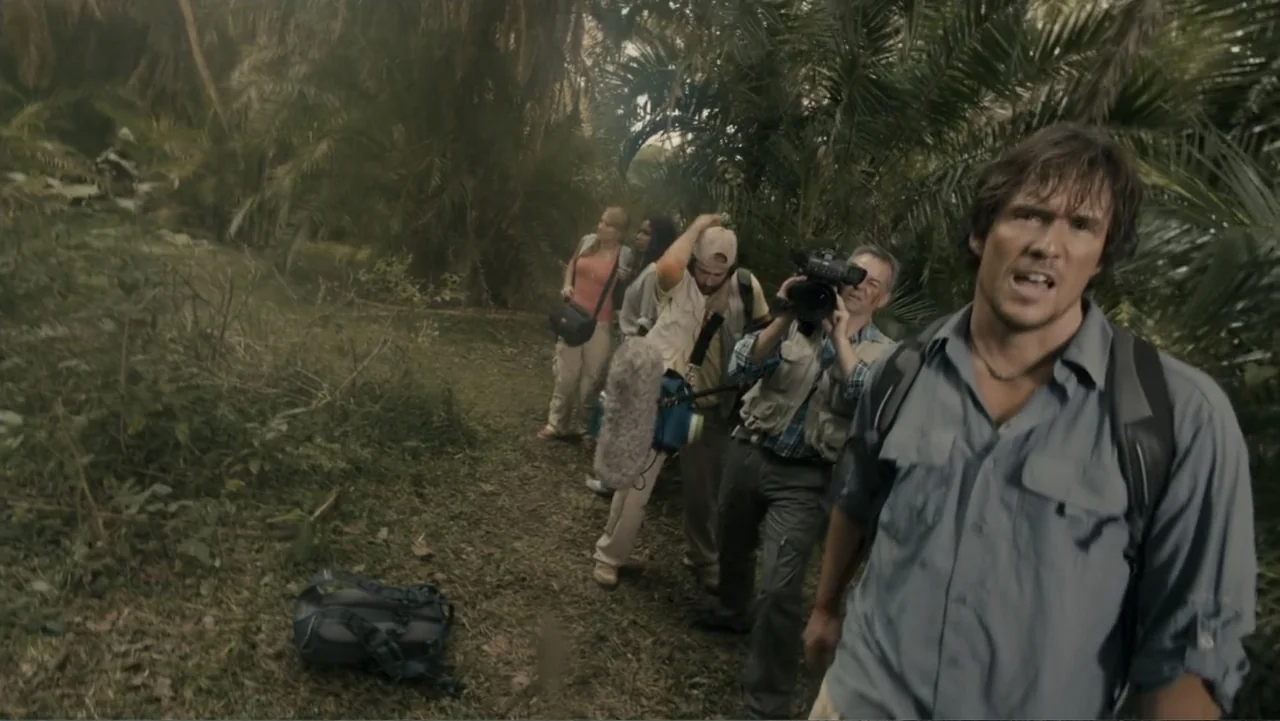 The Dinosaur Project (2012) movie still: medium — A group of people navigate through a dense, green jungle thicket. A man in the foreground speaks…