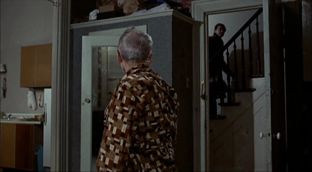 The King Of Marvin Gardens movie still: medium — A residential room featuring a patterned robe-wearing man in the foreground and a staircase. A man…