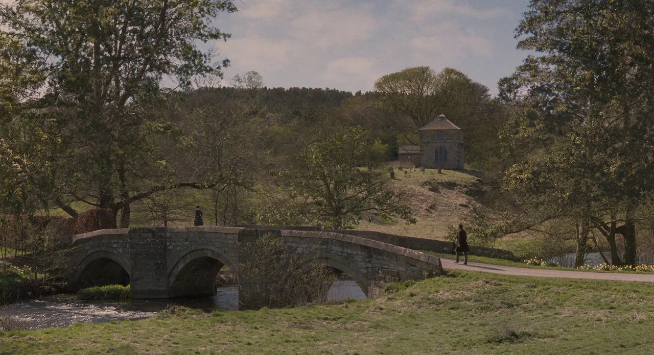 Jane Eyre (2011) movie still: wide — A rural landscape features an old stone arch bridge over a winding river. A small stone building stands atop a…