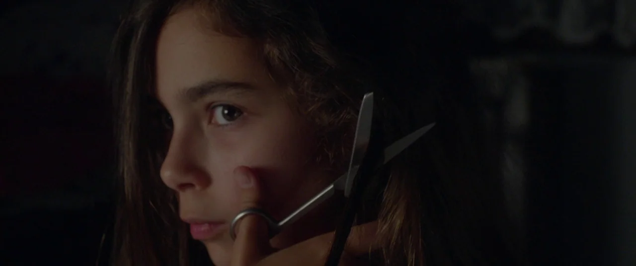 Mustang (2015) movie still: closeup — A dark indoor space provides a low-key background for a close portrait. A young girl holds a pair of metal…
