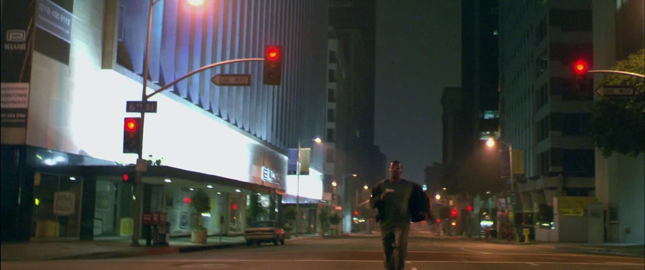 Collateral (2004) movie still: wide — An empty urban intersection at night featuring high-rise buildings and illuminated street signs. A man runs…