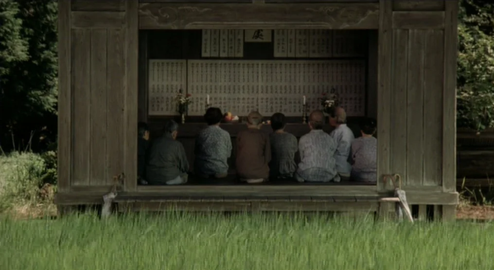 Rhapsody in August (1991) movie still: wide — A wooden open-air structure faces a field of green grass. Several people sit on the floor in front of…