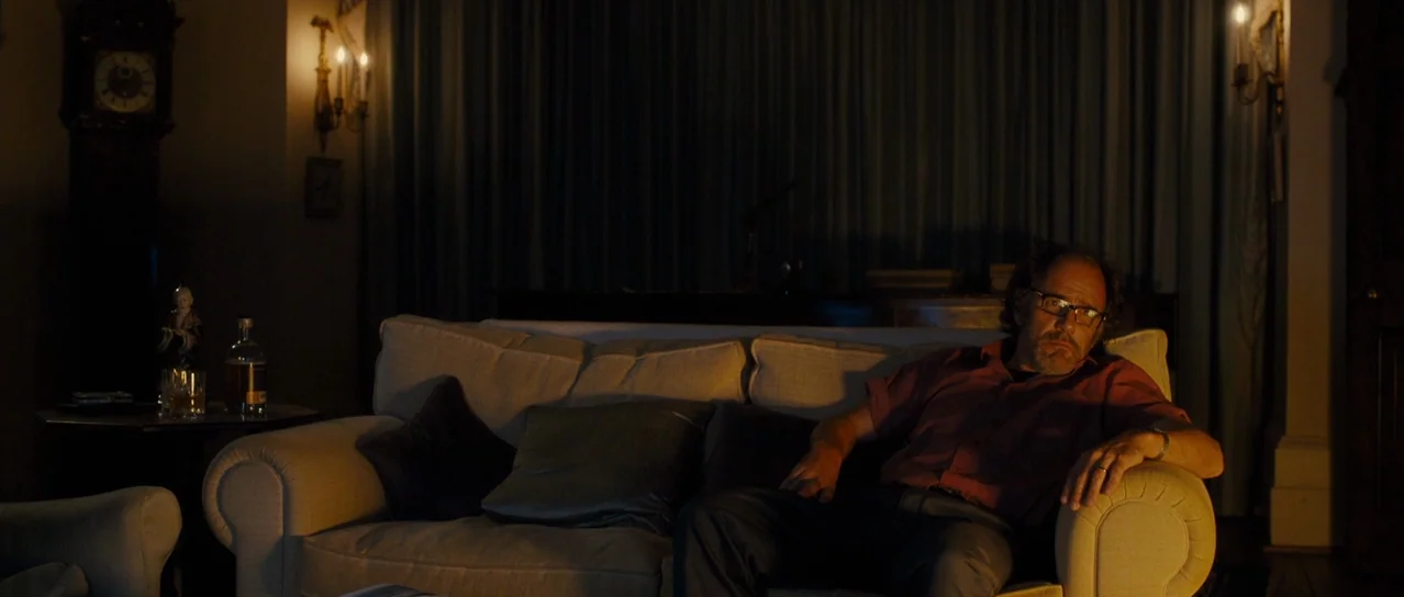 Road Games (2015) movie still: medium — A dimly lit room features a cream sofa positioned against dark floor-to-ceiling curtains. An older man with…