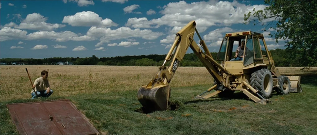 Take Shelter (2011) movie still: wide — An open field under a blue sky with scattered clouds features a construction site. A yellow backhoe…