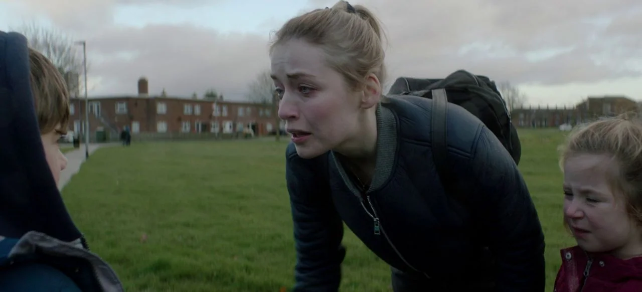 A Good Woman Is Hard To Find (2019) movie still: medium — A grassy field in front of a red brick housing block under overcast skies. A young woman…