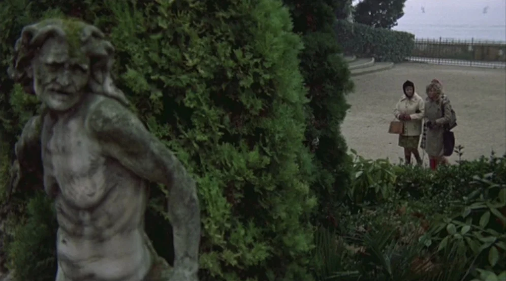 Don’t Look Now (1973) movie still: wide — A public garden features a large weathered stone sculpture in the immediate foreground. Two women in…