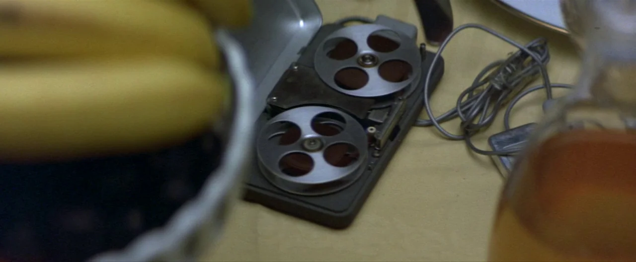Klute (1971) movie still: closeup — A close-up view of a small reel-to-reel tape recorder on a yellow surface. Beside the recorder sit a bowl of…