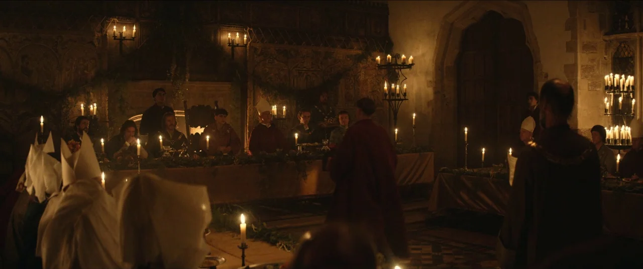 The King movie still: wide — A stone banquet hall features long tables decorated with green garlands and many candles. Several figures wearing…